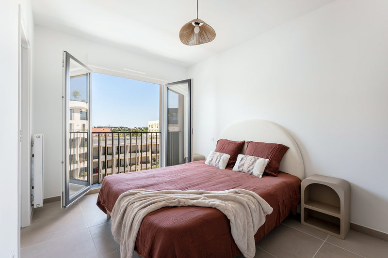 Chambre meublée : tête de lit arrondie, linge terracotta, suspension en corde, vue balcon