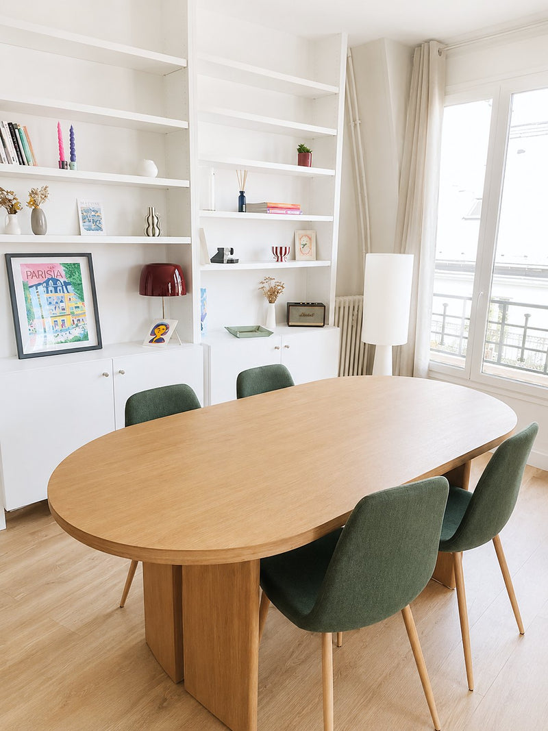 Salle à manger meublée par Les Compositeurs : table ovale en bois, chaises vertes, bibliothèque décorée et lampadaire
